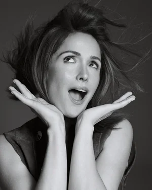 Black and white portrait of Rose Byrne looking upward with hands framing her face and windblown hair against a grey studio backdrop.