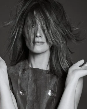 Black and white portrait of Rose Byrne with windblown hair obscuring her eyes, wearing a sleeveless suede buttoned top.