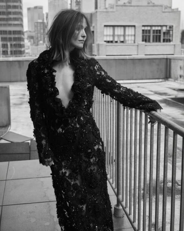 Rose Byrne wearing a long black embellished lace plunging dress while leaning on a metal balcony railing on a city rooftop.