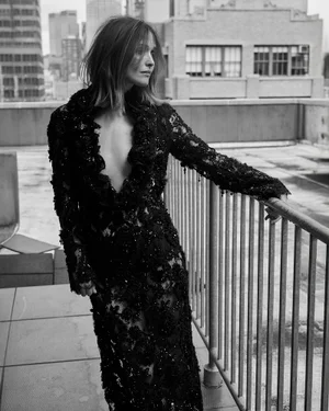 Rose Byrne wearing a long black embellished lace plunging dress while leaning on a metal balcony railing on a city rooftop.