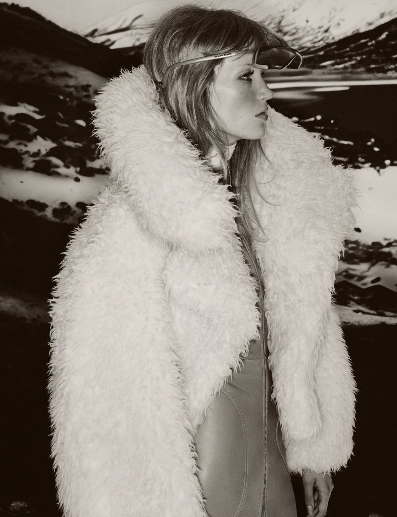 A woman with long hair wearing a white fur coat and sunglasses stands against a blurred background of mountains and water.