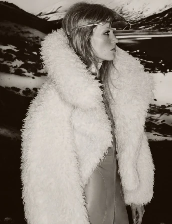 A woman with long hair wearing a white fur coat and sunglasses stands against a blurred background of mountains and water.