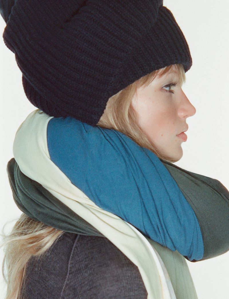 Profile portrait of an adult woman wearing a black knit cap and a blue, green, and white scarf against a white background.