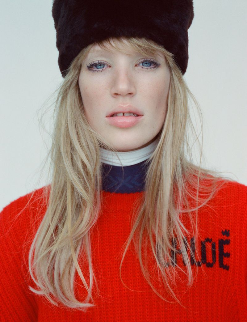 Close-up studio portrait of a fair-skinned woman with blue eyes wearing a black fur hat and red sweater with the word 'CHLOE'.