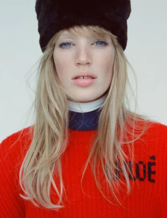 Close-up studio portrait of a fair-skinned woman with blue eyes wearing a black fur hat and red sweater with the word 'CHLOE'.