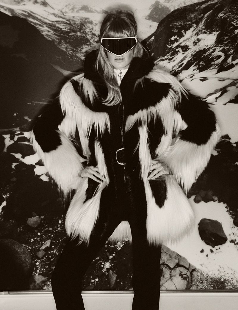 A woman poses in a fur coat and ski goggles against a snowy mountain backdrop, exuding winter fashion and outdoor adventure style.