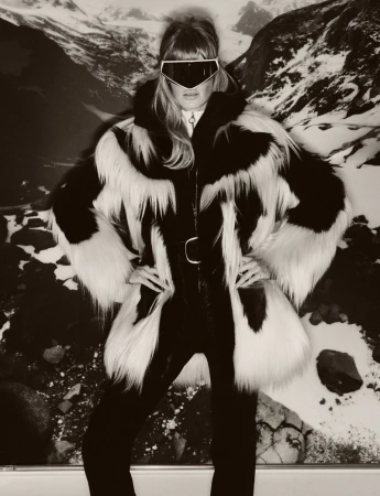 A woman poses in a fur coat and ski goggles against a snowy mountain backdrop, exuding winter fashion and outdoor adventure style.
