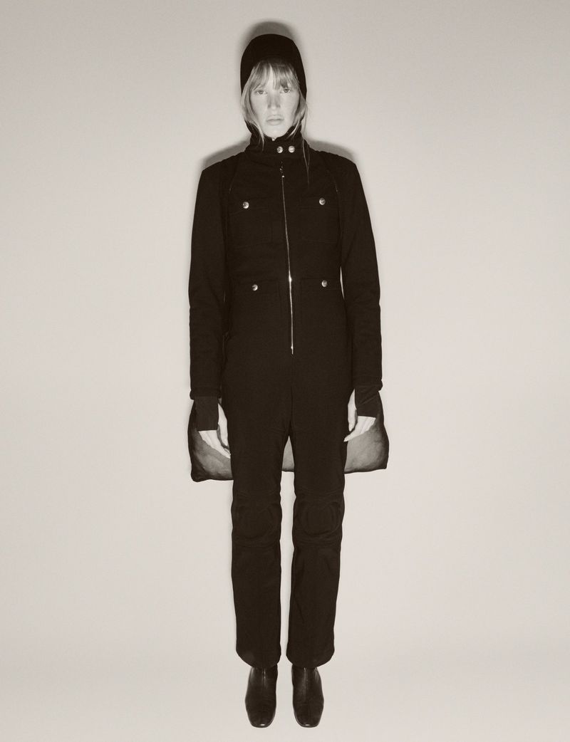 Full-length studio shot of an adult woman in a dark one-piece suit and beanie, standing against a light neutral background with minimal shadows.