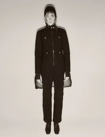 Full-length studio shot of an adult woman in a dark one-piece suit and beanie, standing against a light neutral background with minimal shadows.