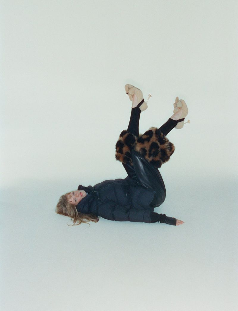 A woman in a black puffer jacket and leopard print leg warmers lies upside down against a white backdrop in a fashion shot.
