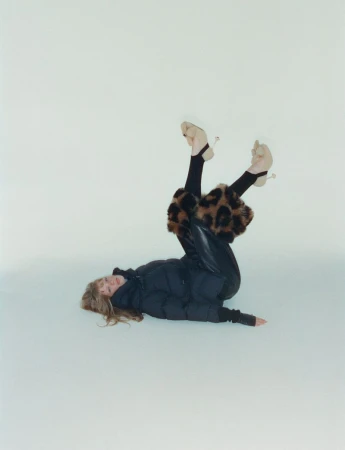 A woman in a black puffer jacket and leopard print leg warmers lies upside down against a white backdrop in a fashion shot.
