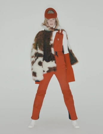 Fashionable woman in orange ski suit, fur coat, and cap poses in a studio setting. A full-length shot with a clean, bright backdrop.