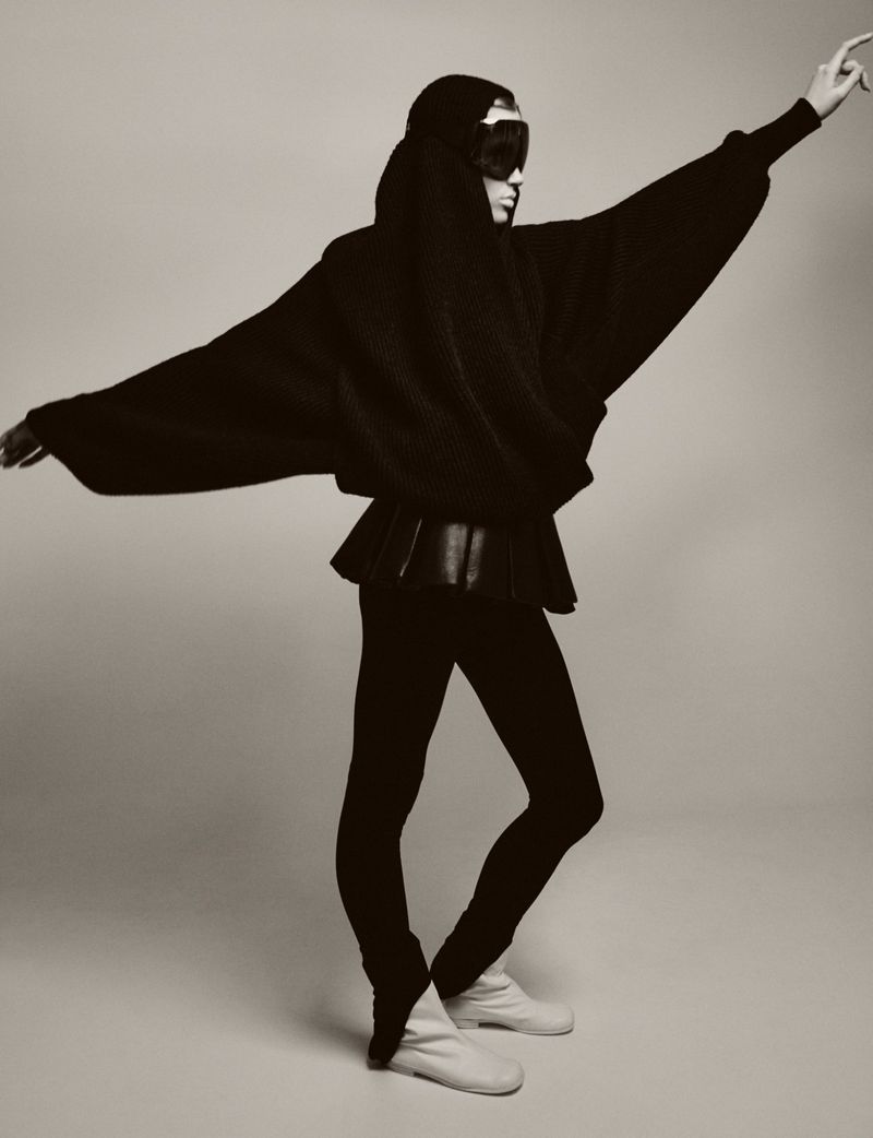 A single person in a studio shot wearing a black hooded sweater, peplum top, leggings, and soft boots striking a dynamic pose in black and white.