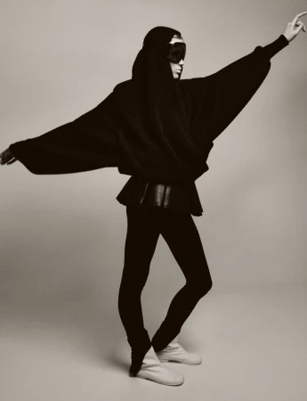 A single person in a studio shot wearing a black hooded sweater, peplum top, leggings, and soft boots striking a dynamic pose in black and white.