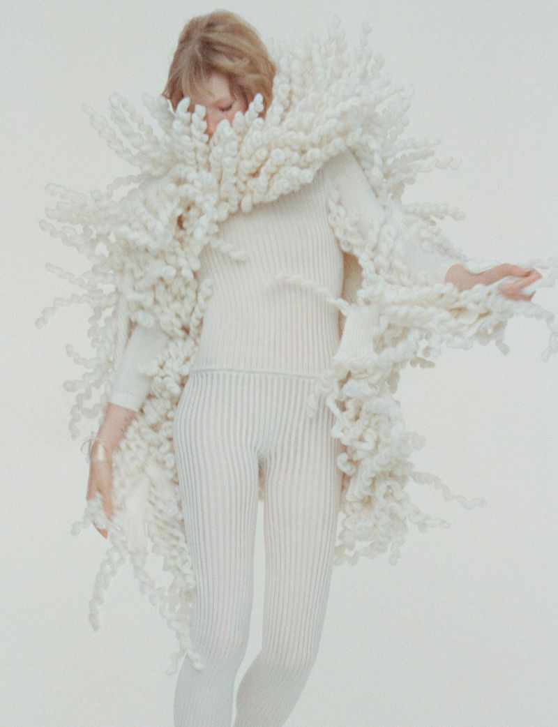 Woman in a full-body ribbed white outfit and large statement piece made of looped white yarn, posing in front of a solid white background.