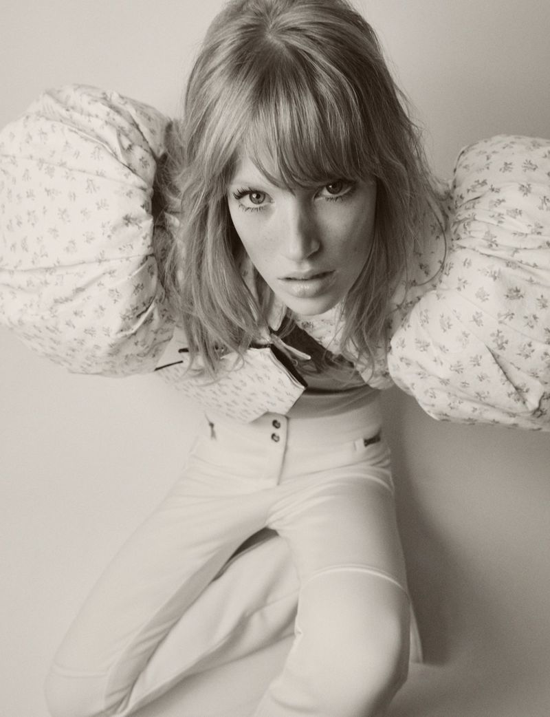 Woman with blonde hair in a patterned top and white pants poses in an eye-level full shot in a photo studio setting.