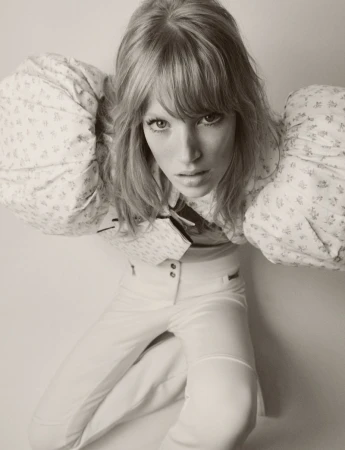 Woman with blonde hair in a patterned top and white pants poses in an eye-level full shot in a photo studio setting.