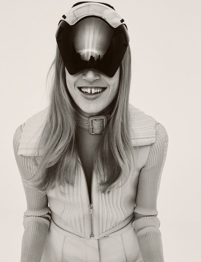 A woman with long blonde hair smiles while wearing a futuristic visor, a beige collared shirt, and a buckled choker in a studio setting.