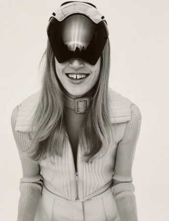 A woman with long blonde hair smiles while wearing a futuristic visor, a beige collared shirt, and a buckled choker in a studio setting.
