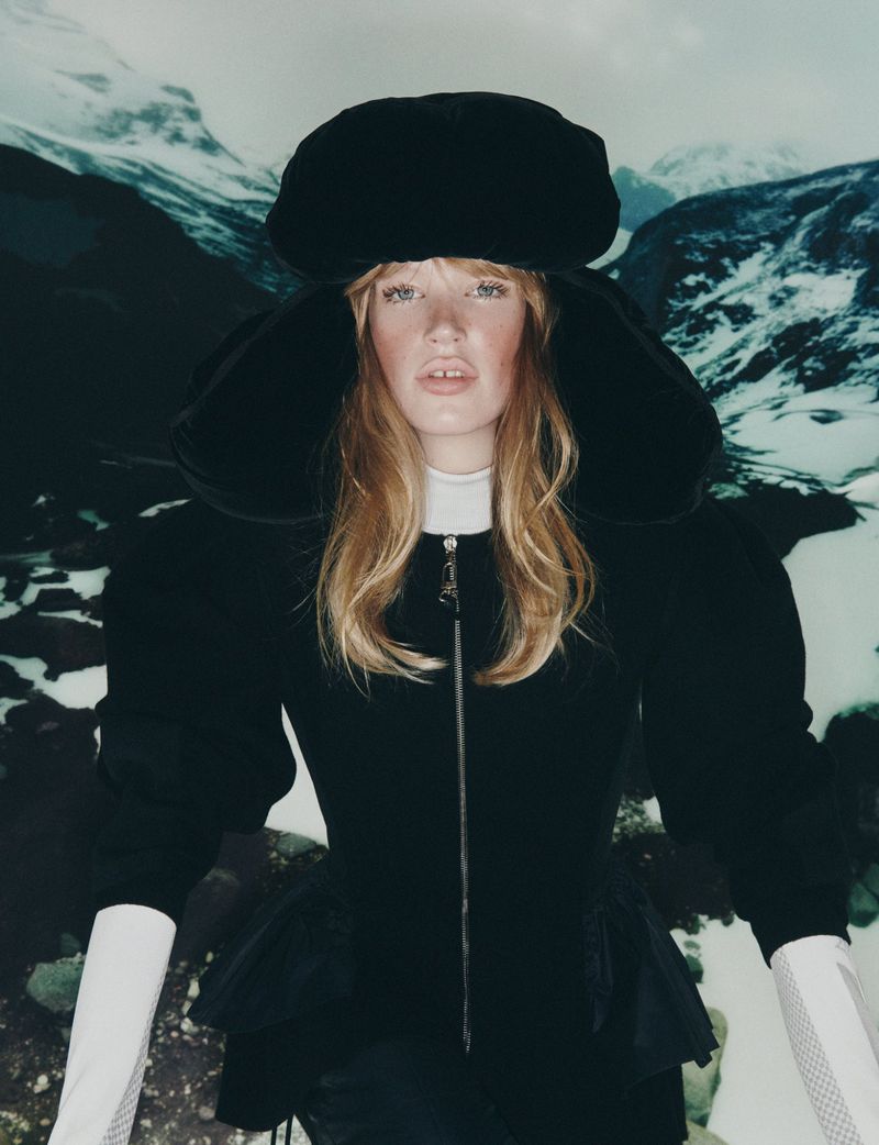 A fair-skinned woman with blonde hair models a black hat and jacket against a snowy mountain backdrop in a fashion editorial.