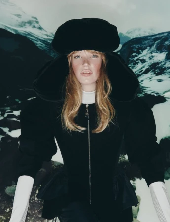 A fair-skinned woman with blonde hair models a black hat and jacket against a snowy mountain backdrop in a fashion editorial.