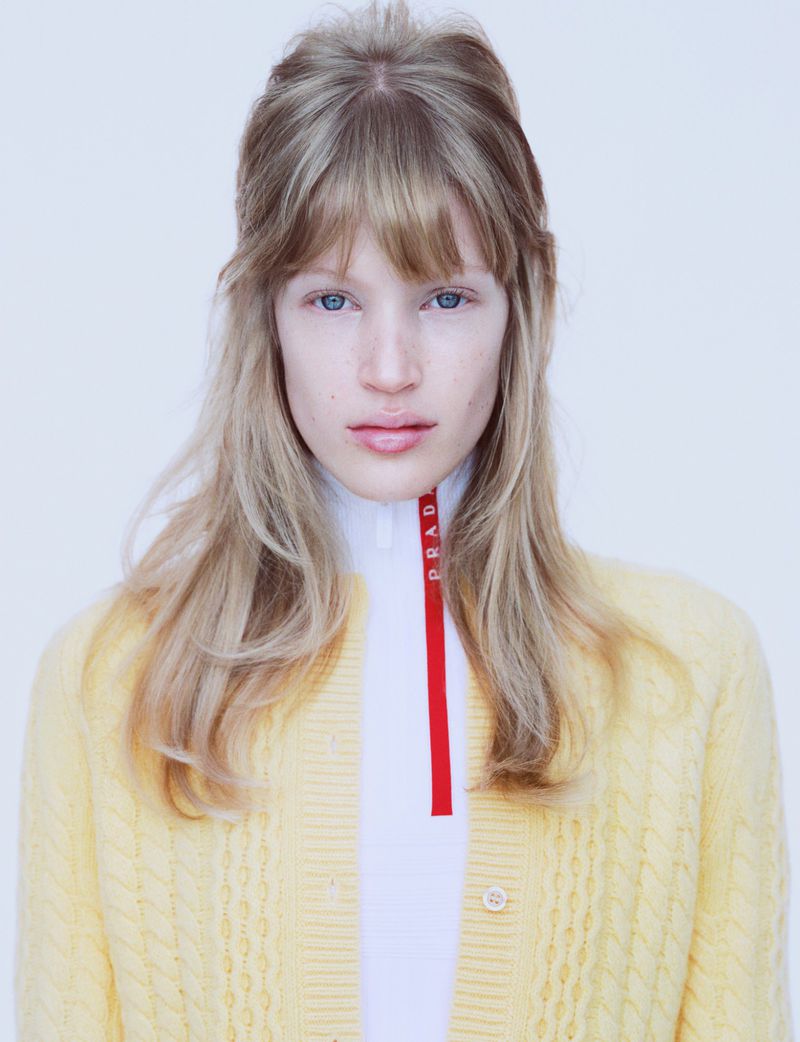 Portrait of a young woman with fair skin and blonde hair wearing a yellow cardigan over a white top with a red Prada logo.