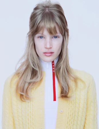 Portrait of a young woman with fair skin and blonde hair wearing a yellow cardigan over a white top with a red Prada logo.