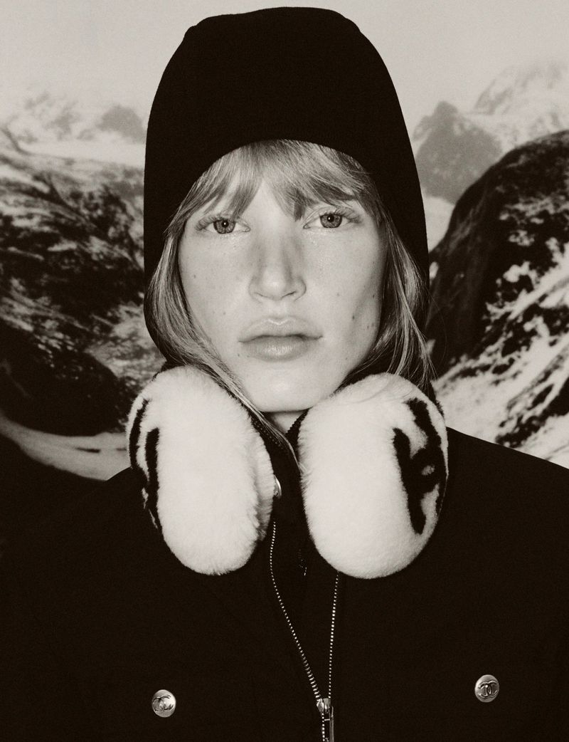 Portrait of a blonde woman wearing a knit cap and fur earmuffs, set against a backdrop of snow-capped mountains in a sepia-toned image.