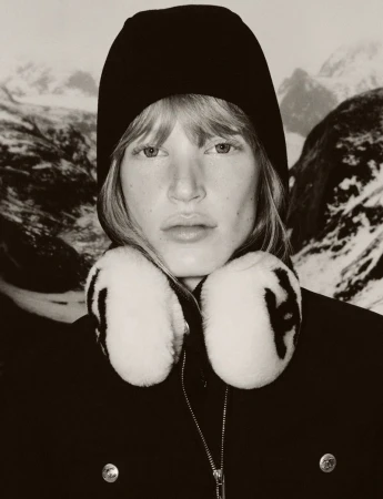 Portrait of a blonde woman wearing a knit cap and fur earmuffs, set against a backdrop of snow-capped mountains in a sepia-toned image.