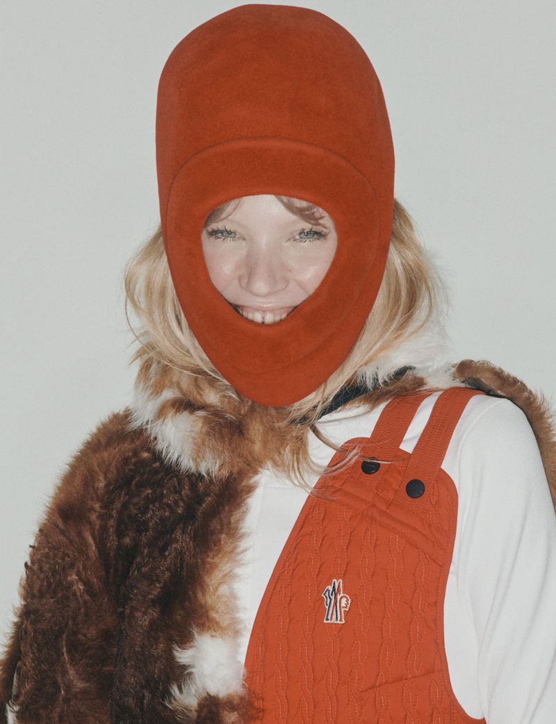 Portrait of a woman with blonde hair wearing an orange balaclava and orange overall dress with fur around her shoulders in a studio.