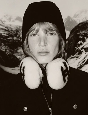 Model wearing a black knit hood and plush white earmuffs over a black jacket in front of a snowy mountain backdrop.