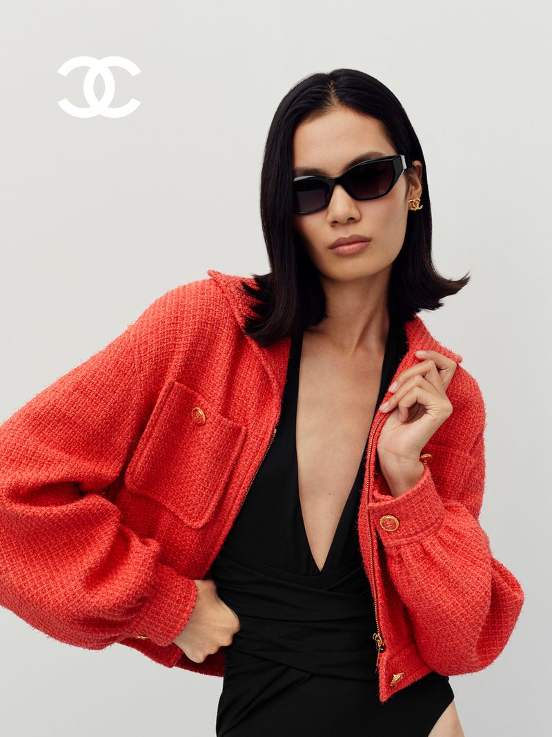 A stylish woman poses indoors wearing a coral tweed jacket, a black bodysuit, and sunglasses, with a Chanel logo in the corner.
