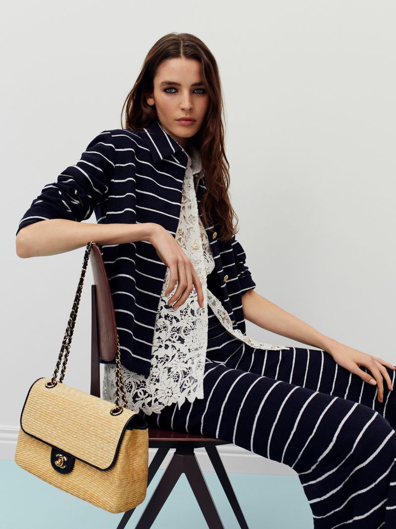 Adult fashion model in a striped suit sitting on a chair in a photo studio with a Chanel handbag, showcasing a commercial look.