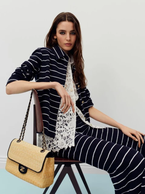 Adult fashion model in a striped suit sitting on a chair in a photo studio with a Chanel handbag, showcasing a commercial look.