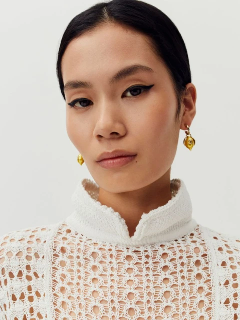 Close-up portrait of an adult female with fair skin, wearing gold earrings, and a white crocheted high-neck top against a white backdrop.