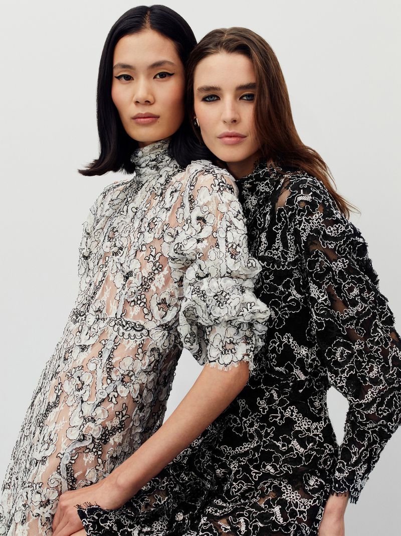 Two women pose closely in elegant black and white lace dresses against a clean backdrop, fashion-forward portrait. Beauty and style.