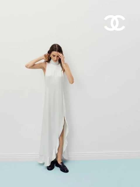A woman poses in a white dress in front of a white wall and turquoise floor. She has one hand on her head.