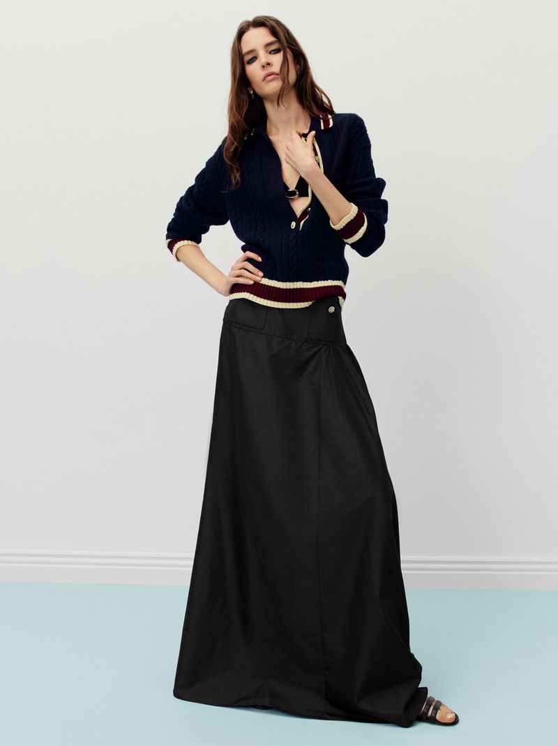 A brunette woman poses in a navy sweater and black maxi skirt against a pale blue wall, showcasing a high-fashion editorial aesthetic.