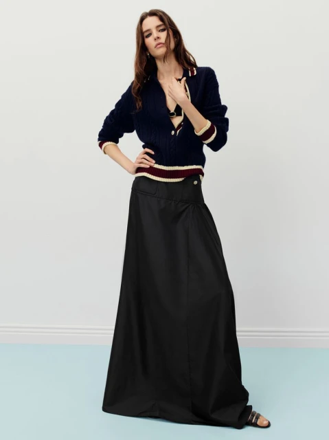 A brunette woman poses in a navy sweater and black maxi skirt against a pale blue wall, showcasing a high-fashion editorial aesthetic.
