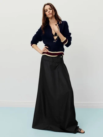 A brunette woman poses in a navy sweater and black maxi skirt against a pale blue wall, showcasing a high-fashion editorial aesthetic.