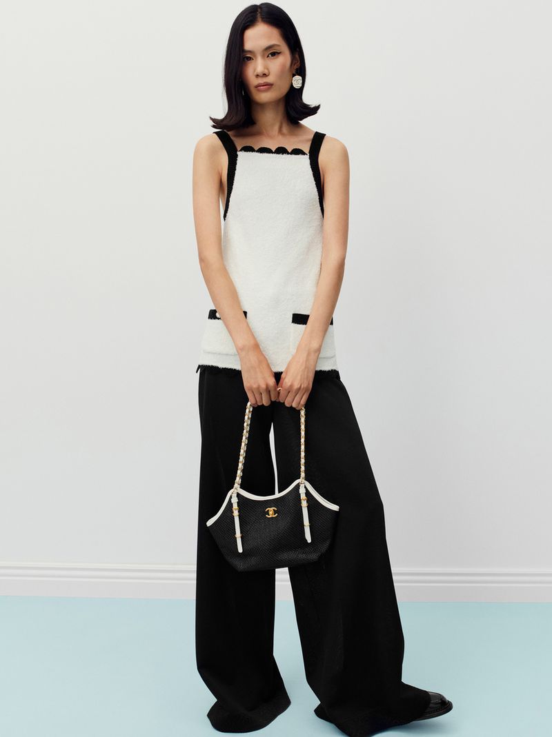 Model wearing white sleeveless top and black trousers holding a chain-strap handbag against a light background