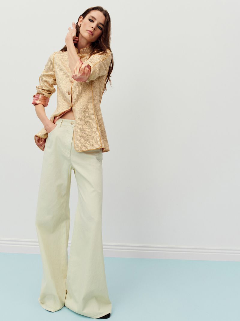 Fashion shot of a woman in a gold blouse and pale green pants posing in front of a white wall with a blue baseboard.