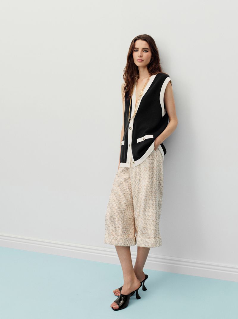 Model wearing a black vest and light patterned culottes posing against a plain wall with a light blue floor.