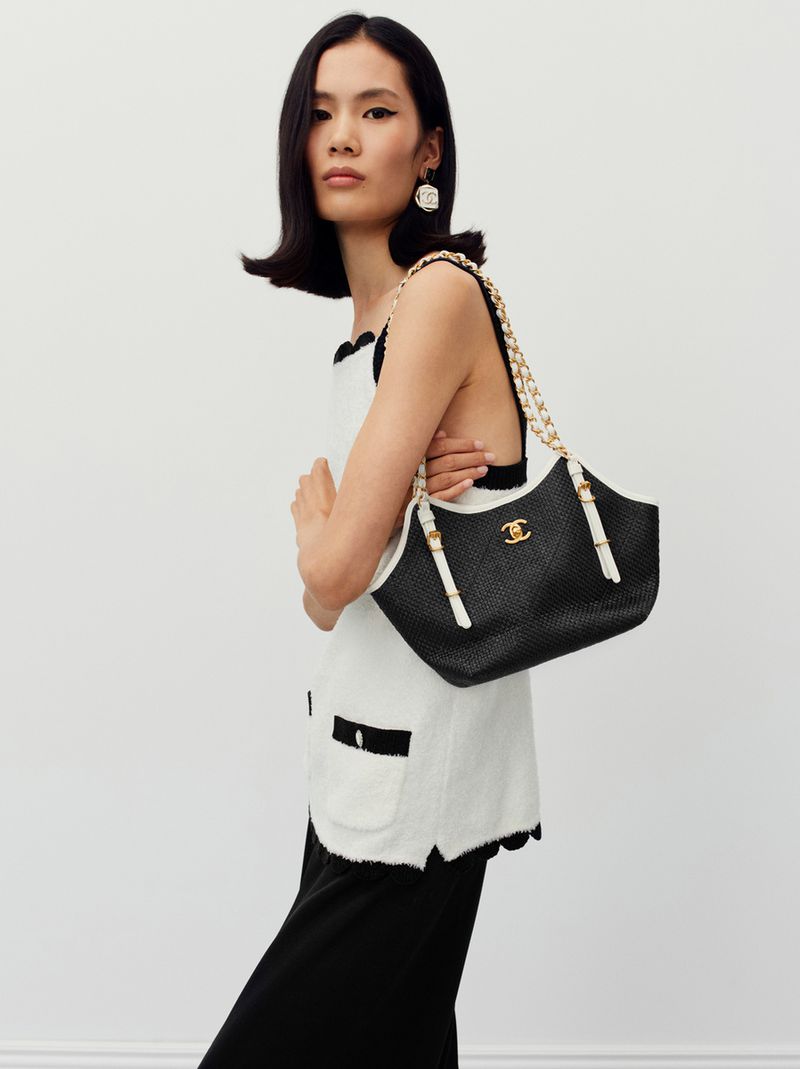 An Asian woman poses in a sleeveless top and skirt, holding a black Chanel handbag with gold chain against a plain backdrop.