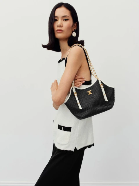 An Asian woman poses in a sleeveless top and skirt, holding a black Chanel handbag with gold chain against a plain backdrop.