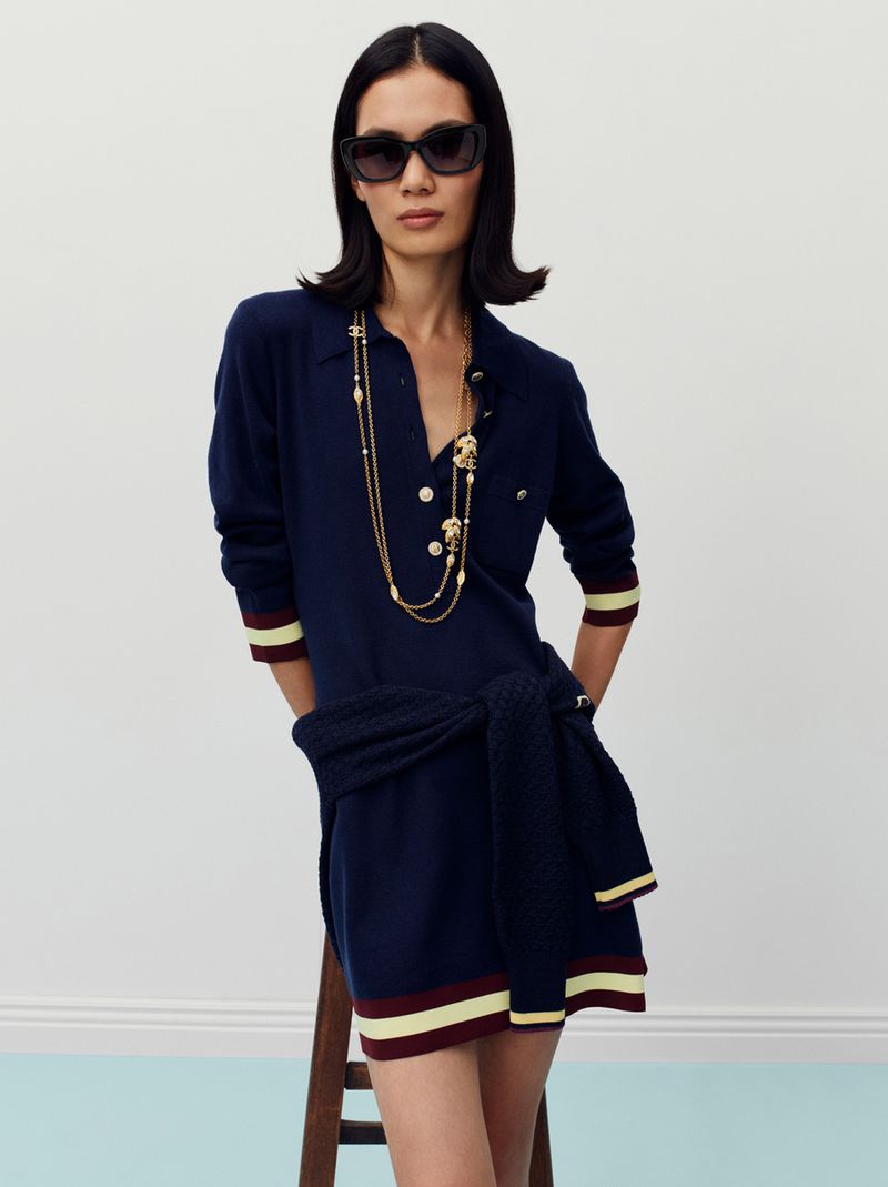 Elegant adult woman with dark hair and sunglasses posing in a navy sweater dress with gold jewelry in a bright indoor studio setting.