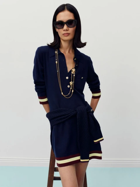 Elegant adult woman with dark hair and sunglasses posing in a navy sweater dress with gold jewelry in a bright indoor studio setting.