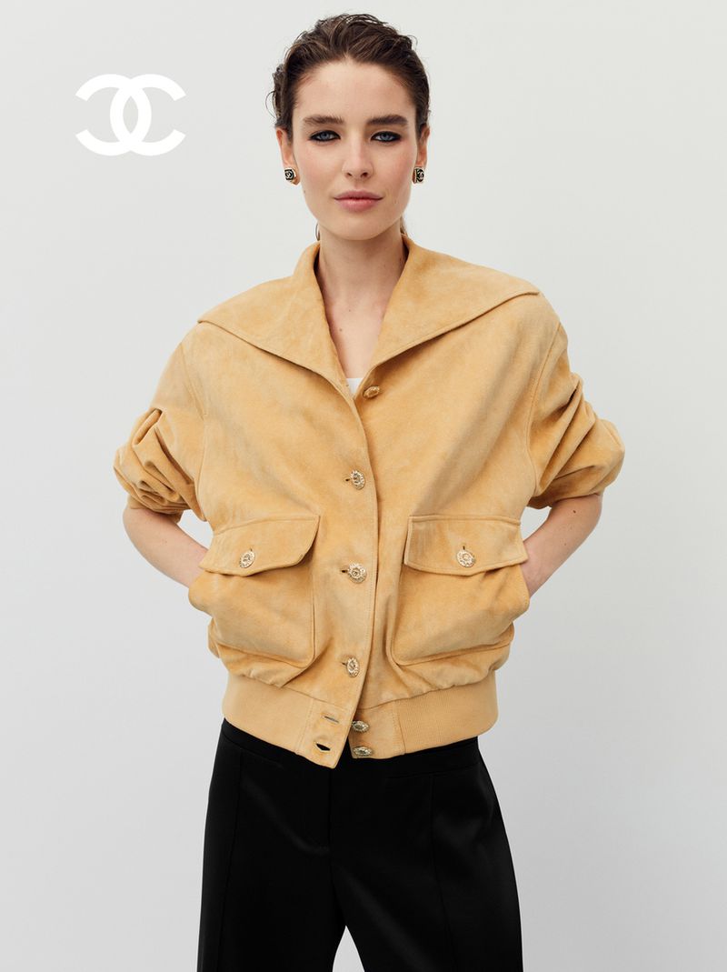 A fair-skinned adult female model wears a tan jacket, black pants, and jewelry in a light-toned indoor setting, with a Chanel logo.