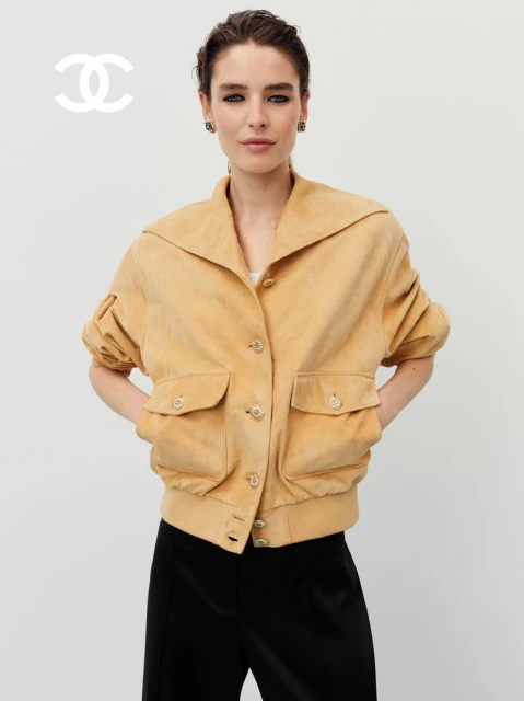 A fair-skinned adult female model wears a tan jacket, black pants, and jewelry in a light-toned indoor setting, with a Chanel logo.