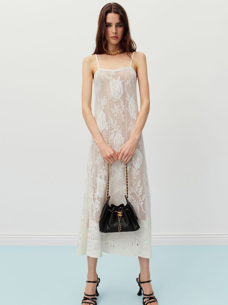 A full shot of a female fashion model showcasing a semi-sheer, rose-patterned dress, a black purse, and black high-heel shoes in a studio setting.